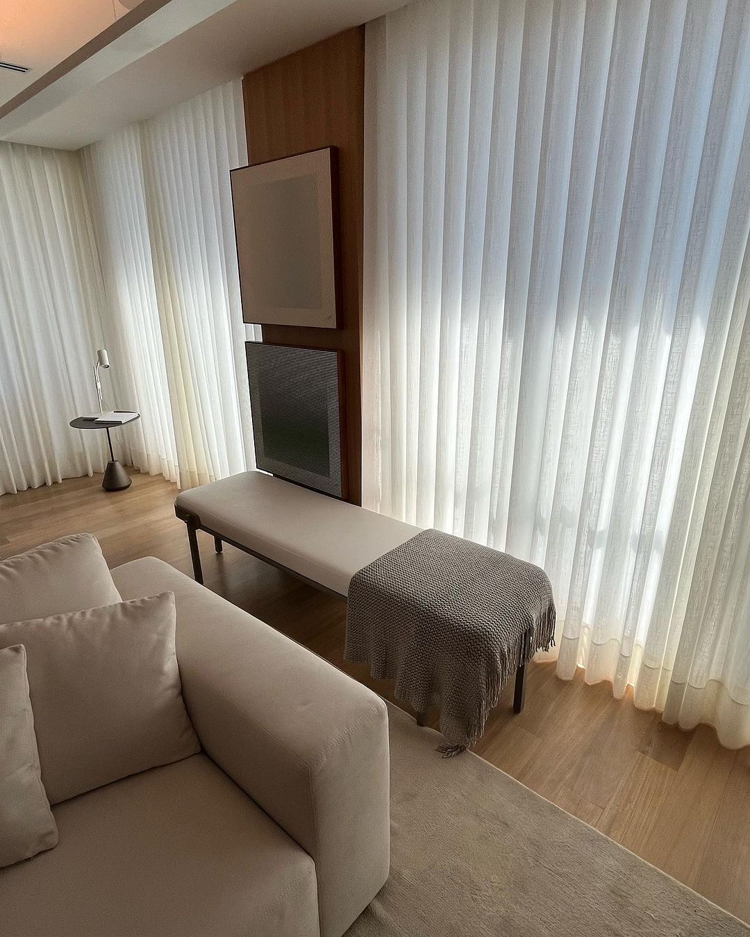 Cortinas brancas em sala com banco e painel de TV
