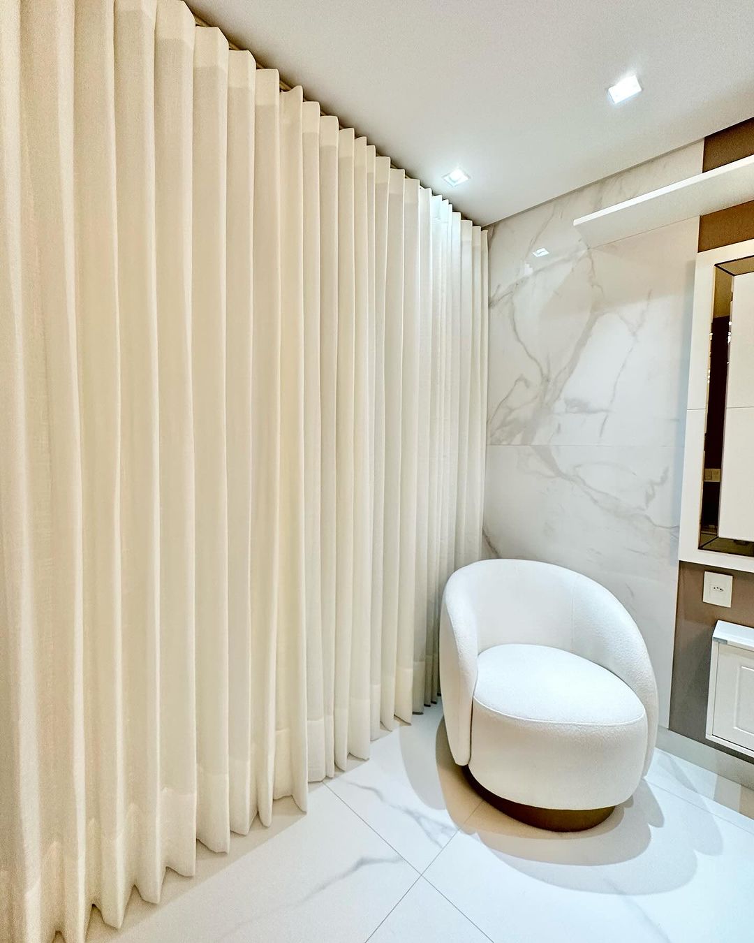 Cortinas plissadas creme com poltrona branca e parede de mármore